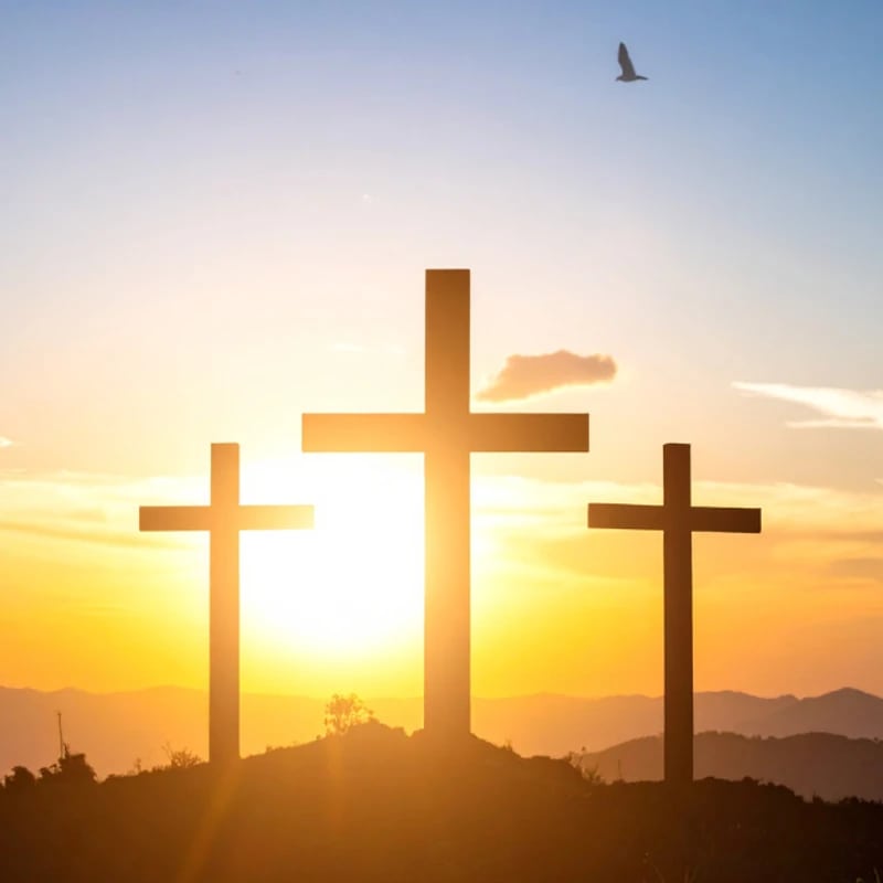 Three wooden crosses are silhouetted on a hilltop against a vibrant sunrise, with mountains in the background and a bird flying in the sky.